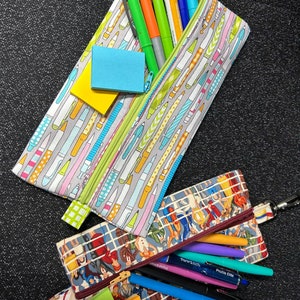 May include: Two zippered pencil cases, one with a colorful pattern of pencils and pens, and the other with a vintage pattern of people. Both cases are filled with pens and pencils, and there are sticky notes in each case.