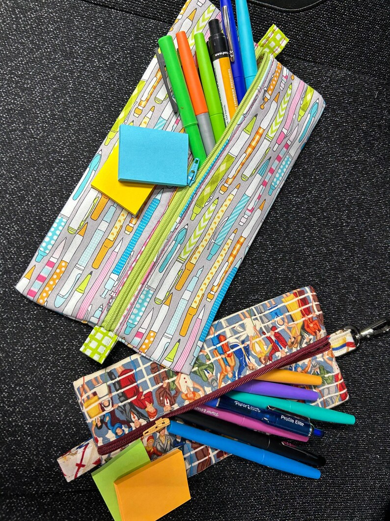 May include: Two zippered pencil cases, one with a colorful pattern of pencils and pens, and the other with a vintage pattern of people. Both cases are filled with pens and pencils, and there are sticky notes in each case.
