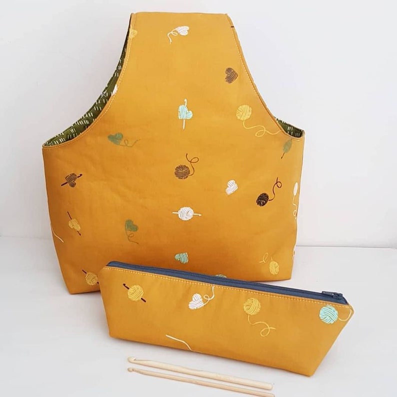 May include: A mustard yellow yarn bag with a zippered pouch and two knitting needles. The bag and pouch have a repeating pattern of yarn balls and hearts.