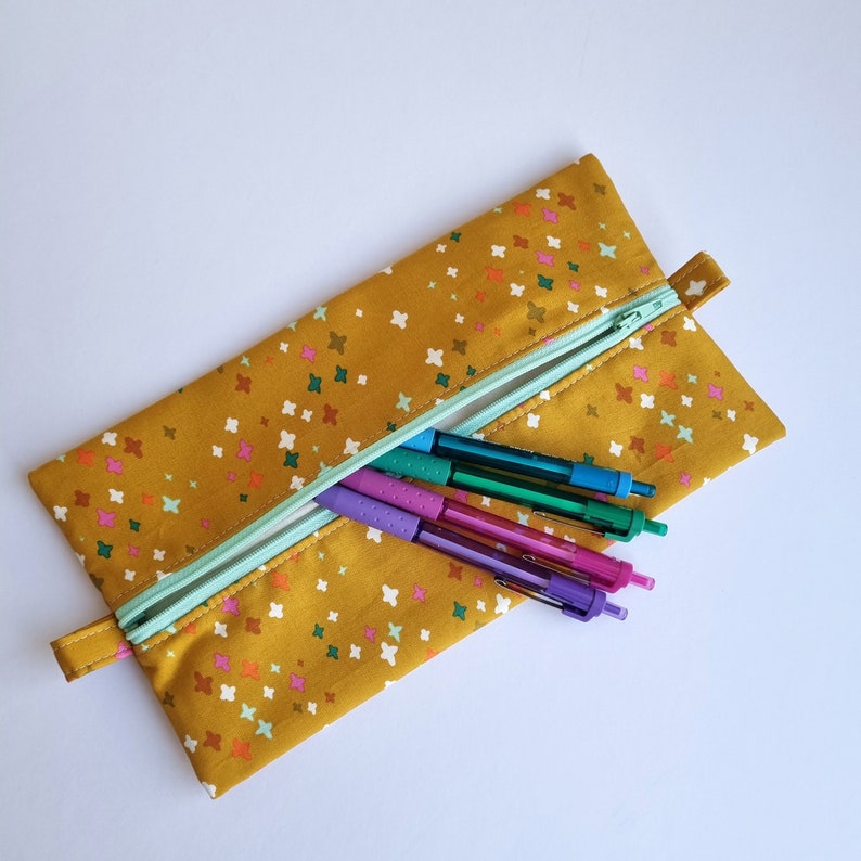 May include: A yellow pencil case with a zipper closure and a colorful pattern of small stars. The case is open and contains five pens in various colors.