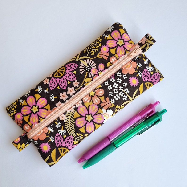 May include: A brown pencil case with a zipper closure and a floral print featuring pink, yellow, and white flowers. The case is lying on a white surface with two pens next to it.
