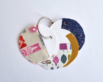 May include: A set of five fabric keyrings with different patterns. The keyrings are attached to a silver ring. The patterns include a cityscape, a floral design, a geometric pattern, a navy blue fabric with white dots, and a mustard yellow fabric.