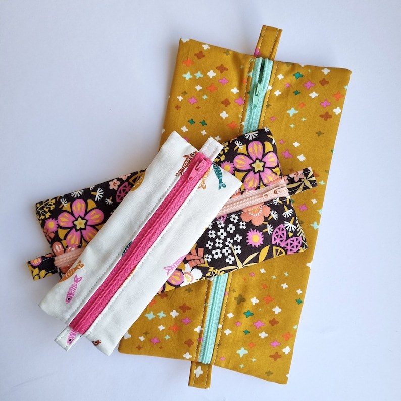May include: Three zippered pouches with floral and fish patterns. The largest pouch is mustard yellow with white stars, the middle pouch is black with pink and white flowers, and the smallest pouch is white with pink and blue fish.