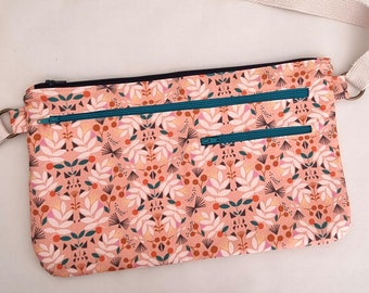Pode incluir: Uma bolsa de ombro de tecido rosa com uma alça branca e dois zíperes turquesa. A bolsa tem um padrão floral com detalhes em laranja, branco e rosa.