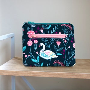 Op de afbeelding: Een zwarte ritszak met een bloemenprint met een witte zwaan, roze en groene bloemen en witte sterren. De tas heeft een roze rits.