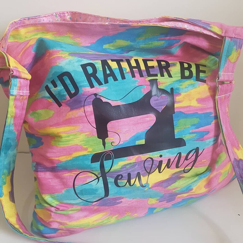 May include: A pink, blue, yellow, and green tote bag with a tie-dye pattern. The bag has a black silhouette of a sewing machine with a heart on it and the text "I'd rather be sewing".