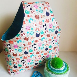 May include: A teal and white fabric tote bag with a cat print. The bag has a wide opening and a handle. The cat print features a variety of cats in different colours and poses.