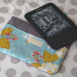 May include: A blue and yellow floral fabric case with a velcro closure for an e-reader. The e-reader is black and has a screen displaying a black and white image. The text on the screen reads "Slide and release the power switch to wake."