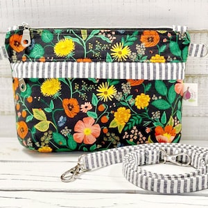 May include: A black zippered pouch with a floral print and a white and black striped strap. The pouch has a zipper closure and a small loop on the side. The strap is adjustable and has a metal clasp.