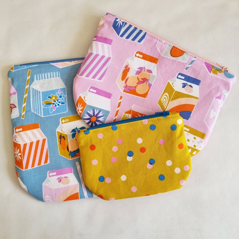 May include: Three fabric pouches with zipper closures. The largest pouch is pink with a white and blue pattern and features a repeated image of a carton of milk with fruit. The medium pouch is blue with a white and yellow pattern and features a repeated image of a carton of milk with fruit. The smallest pouch is yellow with a white and blue polka dot pattern.
