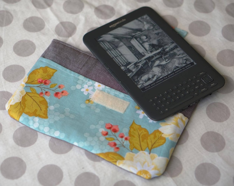 Puede incluir: Una funda de tela floral azul y blanca con un lector electr&oacute;nico negro y gris en la parte superior. El lector electr&oacute;nico tiene una pantalla en blanco y negro con texto que dice "And hold down the power switch to enable".