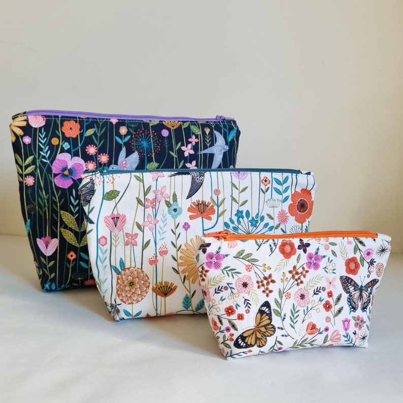 May include: Three floral patterned zippered pouches in different sizes. The largest pouch is black with purple, pink, and white flowers and birds. The medium pouch is white with pink, orange, and yellow flowers. The smallest pouch is white with pink, orange, and yellow flowers and a butterfly.