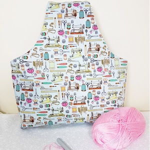 May include: A white fabric tote bag with a repeating pattern of sewing machines, spools of thread, scissors, and other sewing supplies. The bag is filled with pink yarn and a knitting needle.