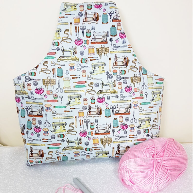 May include: A white fabric tote bag with a repeating pattern of sewing machines, spools of thread, scissors, and other sewing supplies. The bag is filled with pink yarn and a knitting needle.