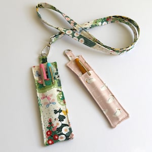 May include: Two fabric pen holders with a floral and swan print. The holders are attached to a white and green floral lanyard. The holders are designed to hold pens or pencils.