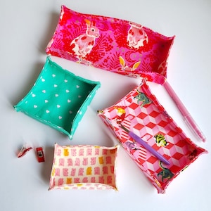 May include: Four fabric storage baskets in different patterns and colours. The baskets are made of a soft, quilted fabric and have a square or rectangular shape. The patterns include pink bunnies, green hearts, pink and white geometric shapes, and pink and yellow bears.