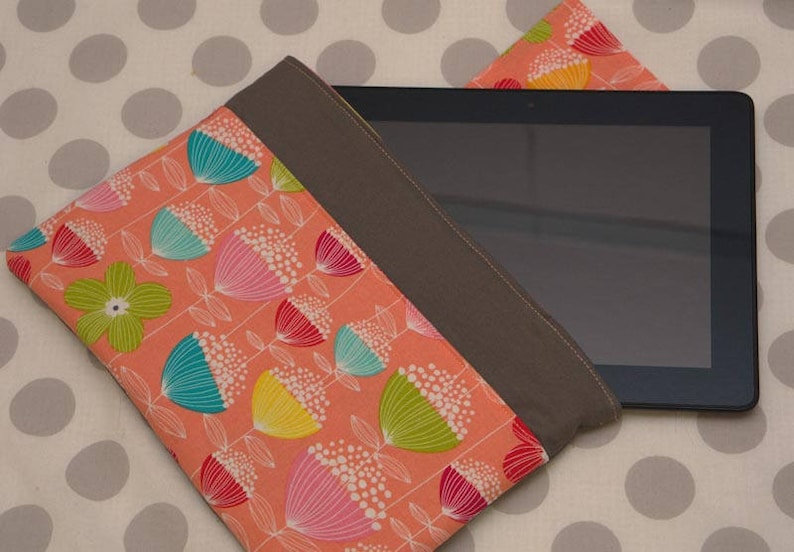 Puede incluir: Una funda para tablet con una solapa de tela marr&oacute;n y un estampado floral. La funda est&aacute; hecha de una tela suave de color melocot&oacute;n con un patr&oacute;n repetitivo de flores estilizadas en tonos de rosa, amarillo, azul y verde.