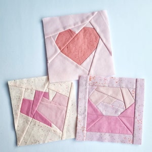 May include: Three pink and white fabric squares with a heart, a swirl, and a diamond pattern. The fabric squares are made with a patchwork technique.