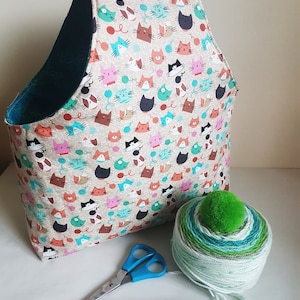 May include: A teal and beige fabric tote bag with a cat print. The bag is filled with yarn and has a green pom-pom on top. There are also knitting needles and scissors on the table.