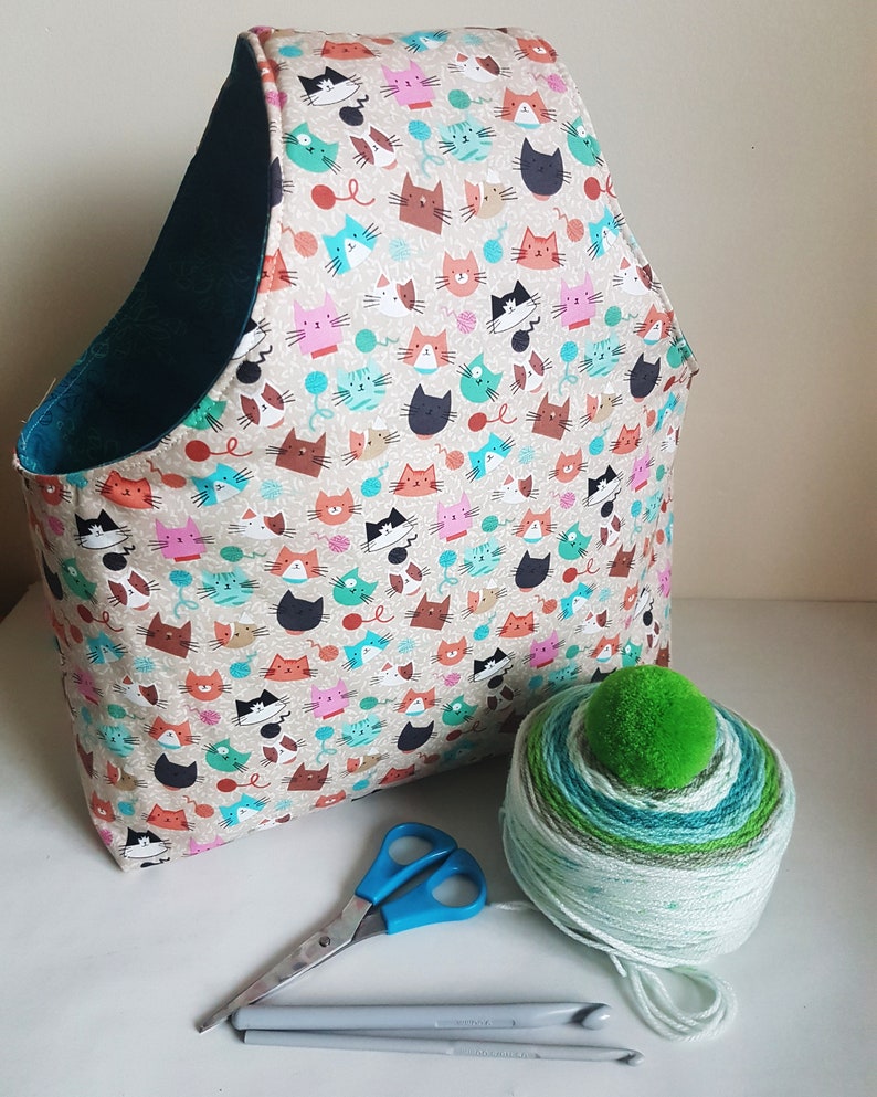 May include: A teal and beige fabric tote bag with a cat print. The bag is filled with yarn and has a green pom-pom on top. There are also knitting needles and scissors on the table.