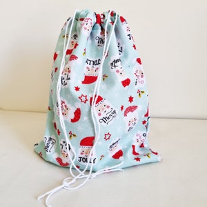 May include: A light blue drawstring bag with a white drawstring. The bag is decorated with a Christmas pattern featuring Santa Claus, snowflakes, and the words "Merry" and "Jolly".