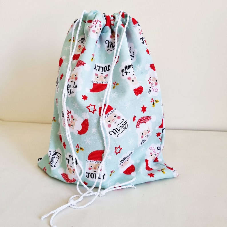 May include: A light blue drawstring bag with a white drawstring. The bag is decorated with a Christmas pattern featuring Santa Claus, snowflakes, and the words "Merry" and "Jolly".