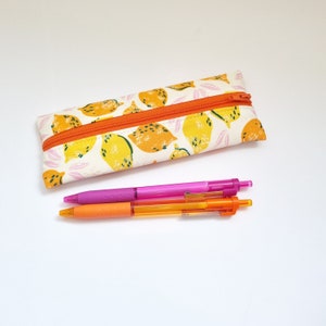 May include: A white pencil case with a zipper closure and a lemon print design. The case is filled with two pens, one pink and one orange.