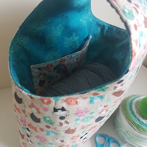 May include: A teal fabric tote bag with a cat print design. The bag has a pocket on the inside and is filled with yarn. A ball of yarn and a pair of blue scissors are on the table next to the bag.