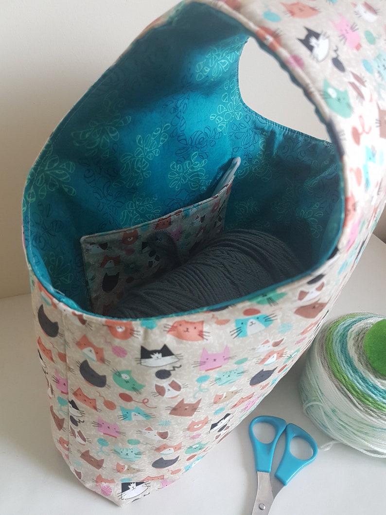 May include: A teal fabric tote bag with a cat print design. The bag has a pocket on the inside and is filled with yarn. A ball of yarn and a pair of blue scissors are on the table next to the bag.