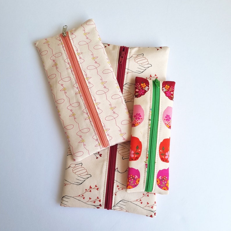May include: Three zippered pouches with different patterns. The largest pouch is cream with a pink and yellow pattern of lines and hearts. The middle pouch is cream with a red and pink pattern of hands and branches. The smallest pouch is cream with a red, pink, and white pattern of flowers.