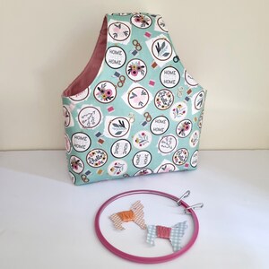 May include: A teal fabric tote bag with a pink lining and a floral print featuring the text "Home Sweet Home" and "Hello". The bag is sitting on a white surface next to a pink embroidery hoop with two pieces of fabric inside.