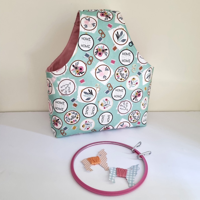 May include: A teal fabric tote bag with a pink lining and a floral print featuring the text "Home Sweet Home" and "Hello". The bag is sitting on a white surface next to a pink embroidery hoop with two pieces of fabric inside.