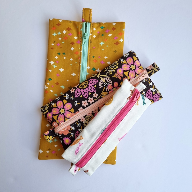 May include: Three fabric pencil cases with zippers. The largest case is mustard yellow with white stars. The middle case is black with pink and white flowers. The smallest case is white with pink and blue fish.