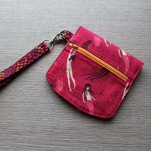 May include: A pink fabric pouch with a zipper closure and a matching lanyard. The pouch features a repeating pattern of mermaids and sea creatures.