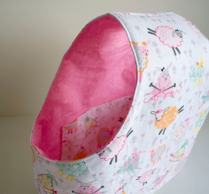 May include: A pink and white fabric storage bin with a sheep pattern. The bin is lined with pink fabric and has a rounded shape.