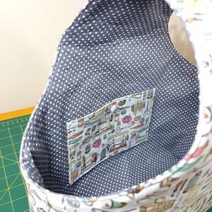 May include: A white fabric tote bag with a gray polka dot lining. The bag has a small pocket on the inside with a sewing themed print. The bag is empty.