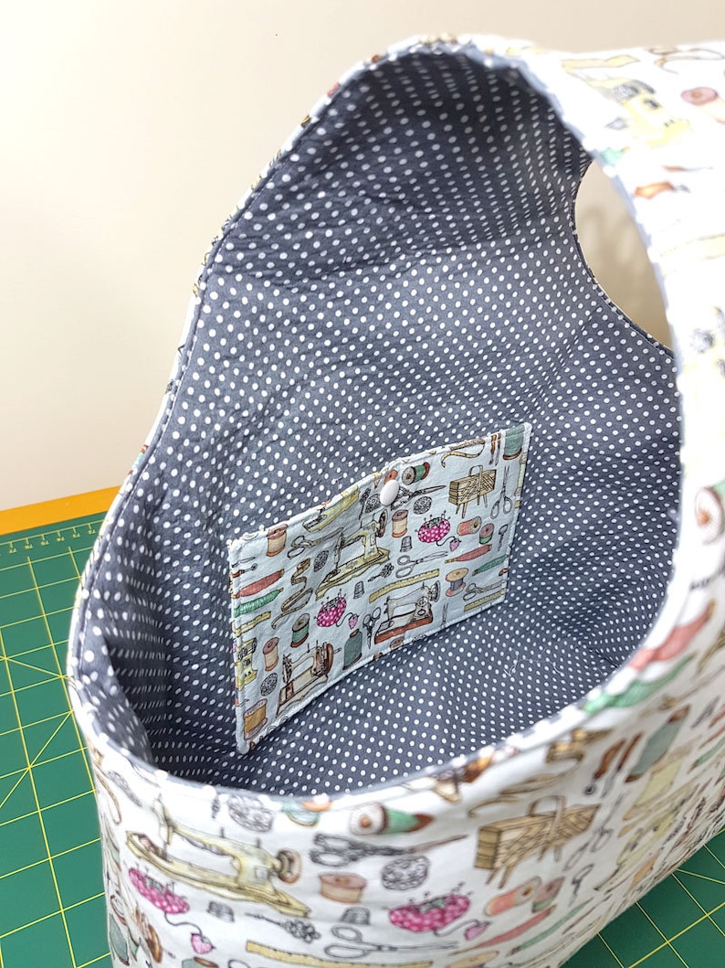 May include: A white fabric tote bag with a gray polka dot lining. The bag has a small pocket on the inside with a sewing themed print. The bag is empty.