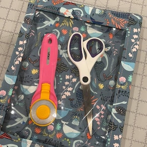 Puede incluir: Una bolsa de tela azul con una ventana de plástico transparente, con un estampado de sirenas y ballenas. La bolsa contiene un cortador rotatorio rosa y un par de tijeras blancas.