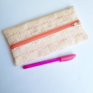 May include: A beige pencil case with a zipper closure. The case is decorated with a repeating pattern of pink and gold "love" lettering. A pink pen is lying next to the case.