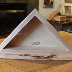 May include: A wooden triangular shadow box with a glass front. The box has a light wood finish and a dark wood base. The box is labeled "Woodcrafted by Joshua Chilson".
