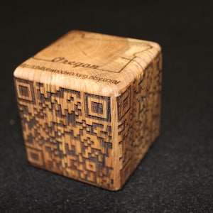 May include: A wooden cube with a laser-engraved QR code and the word "Oregon" on the top. The cube is made of light brown wood with a natural grain pattern.