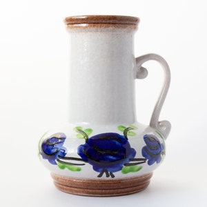 May include: A white ceramic vase with a brown rim and handle. The vase has a floral design with blue and green flowers.