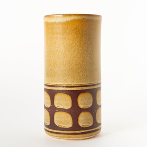 May include: A tall, cylindrical ceramic vase with a brown and yellow geometric pattern. The vase is a light brown color with a darker brown band around the middle.