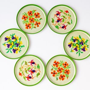May include: Set of six green and white coasters with embroidered floral designs. Each coaster features a different floral pattern with various colors, including orange, yellow, purple, and green.