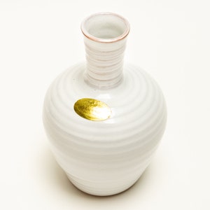 May include: White ceramic vase with a gold oval sticker. The vase has a wide base and a narrow neck. The vase is decorated with a swirling pattern.