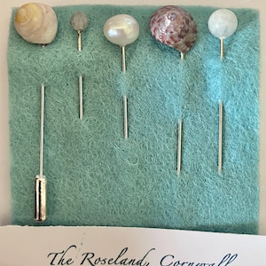 Könnte beinhalten: Eine Sammlung dekorativer Anstecknadeln mit natürlichen Elementen wie Muscheln und Perlen, präsentiert auf einem türkisfarbenen Filzhintergrund. Die Nadeln haben silberne Stiele. Der Text "The Roseland, Cornwall" ist unten zu sehen.