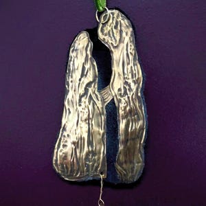 Könnte beinhalten: Handgefertigter Metallschmuck mit zwei strukturierten Figuren, verbunden durch eine dunkelblaue Rückseite. Ein kleiner Sternanhänger hängt unten, mit einem grünen Band zum Aufhängen. Einzigartiges, künstlerisches Design.