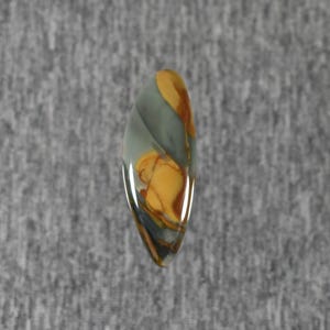 May include: A polished, teardrop-shaped gemstone with a mix of colors. The stone features swirls of gray, tan, and brown. The gemstone is set against a gray background.