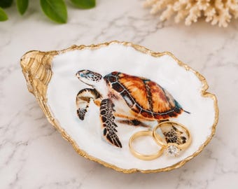 May include: A decorative oyster shell trinket dish featuring a hand-painted sea turtle on the interior. The shell has a smooth white surface with a metallic gold-painted rim. Two gold-toned rings, one with a clear stone, rest inside the dish.