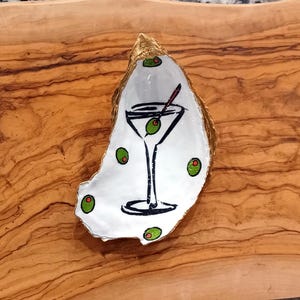 May include: A white oyster shell with gold accents, painted with a martini glass and green olives. The shell is on a wooden surface.